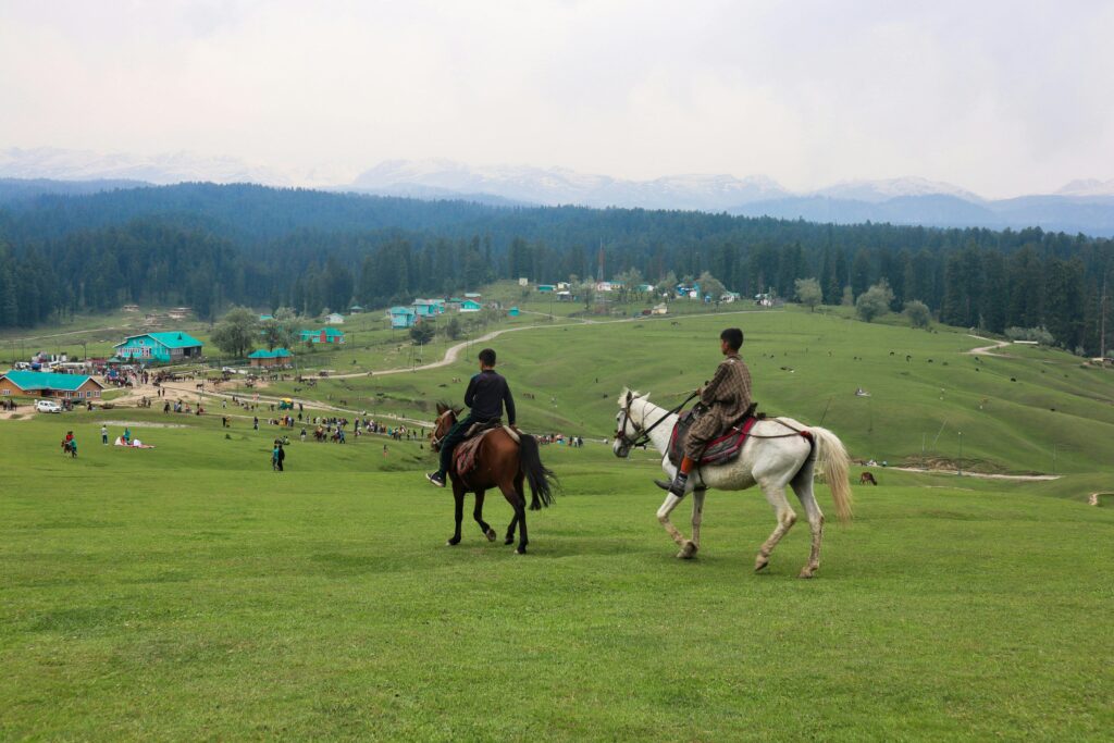 Two horseback riders traverse the lush, rolling hills of Yousmarg with stunning mountain views.