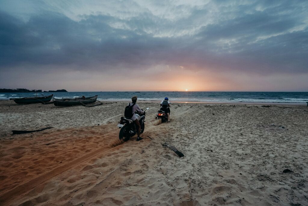 Two motorcyclists enjoying a scenic beach ride at sunset with ocean views and colorful sky.