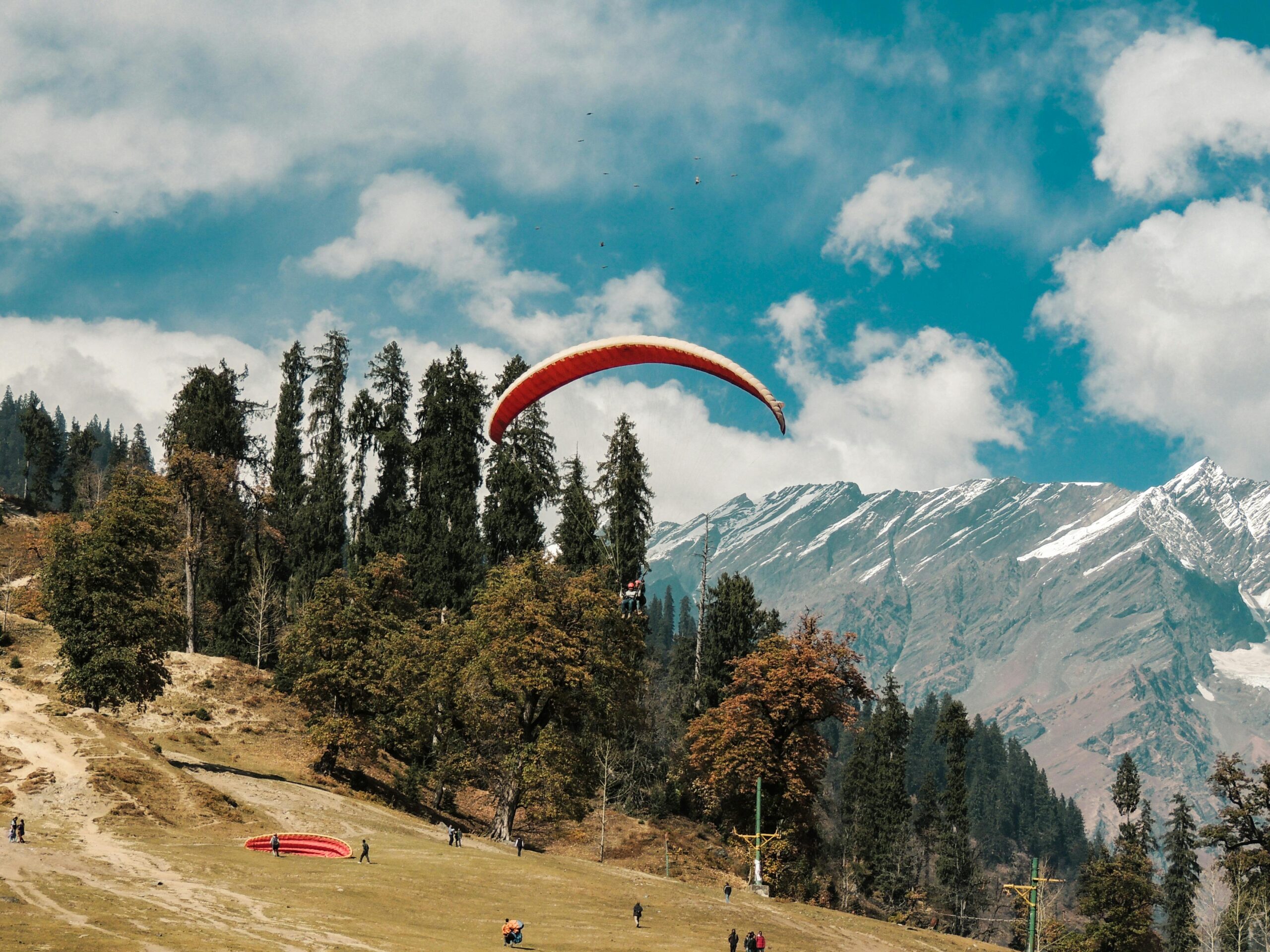 A thrilling paragliding scene over a mountainous landscape with lush forests and snowy peaks.